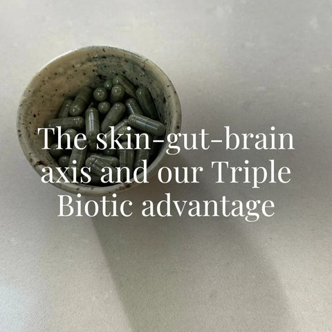 A ceramic bowl filled with greenish-black supplement capsules sits on a clean cream-colored surface. The image is simple, functional, and natural. The overlaid text reads: “The skin-gut-brain axis and our Triple Biotic advantage.”