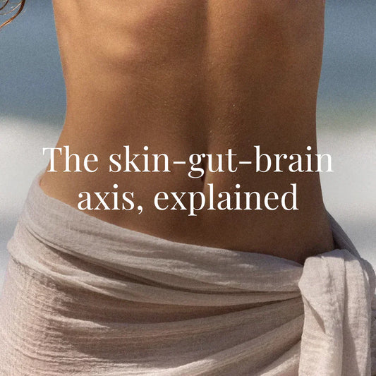 A woman’s toned back and waist, wrapped in a light gauze wrap, faces the ocean. The focus is on the midsection, emphasizing gut health. White serif text reads: “The skin-gut-brain axis, explained.”